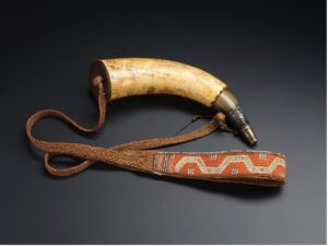 An 18th-century powder horn likely made from a hollowed-out animal horn, featuring a curved shape with a plug at the wide end and a spout at the narrow tip. The surface is engraved with decorative motifs and the strap is either Haudenosaunee or Huron-Wendat.