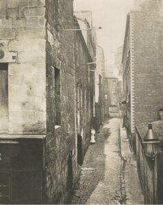 Black and white photograph of a narrow alleyway between brick buildings with a narrow brick lane running between.